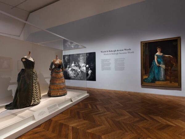 Dwie suknie wieczorowe z epoki w odcieniach zieleni i brązu eksponowane w muzeum. Obok nich znajduje się duży obraz przedstawiający damę w turkusowej sukni, a na ścianie tekst opisujący przekształcenie firmy Worth & Bobergh w dom mody Worth. To zdjęcie z "Worth: Inventing haute couture" w Petit Palais w Paryżu.