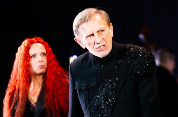 Dwójka aktorów w intensywnej scenie teatralnej — mężczyzna w czarnym kostiumie z błyszczącym elementem na ramieniu wyraża silne emocje, podczas gdy kobieta o długich, rudych, kręconych włosach patrzy na niego z powagą. Kadr ze sztuki "Król Lear" Grzegorza Wiśniewskiego.