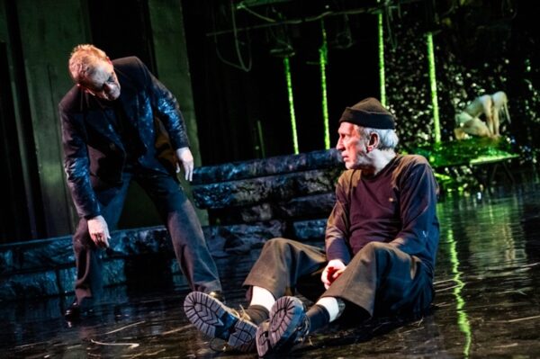 Scena teatralna z dwoma aktorami — jeden z nich stoi pochylony w stronę drugiego, który siedzi na ziemi w czarnym stroju i czapce, na tle mrocznej, industrialnej scenografii. Kadr ze sztuki "Król Lear" Grzegorza Wiśniewskiego.