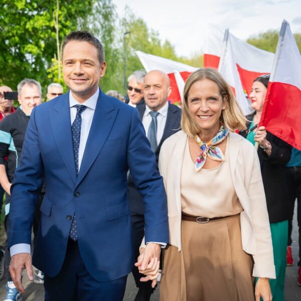 Jest to Rafał Trzaskowski, kandydat na Prezydenta Polski w 2025 roku, wraz z żoną Małgorzata Trzaskowską, która reprezentuje styl Pierwszych Dam. Mężczyzna w niebieskim garniturze i kobieta w jasnym stroju z kolorową apaszką idą razem, trzymając się za ręce; wokół nich tłum ludzi z biało-czerwonymi flagami.