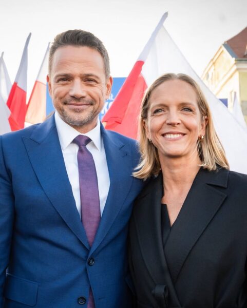 Mężczyzna w niebieskim garniturze i kobieta w czarnym płaszczu stoją razem, uśmiechają się do aparatu, w tle widoczne są biało-czerwone flagi. Jest to Rafał Trzaskowski, kandydat na Prezydenta Polski w 2025 roku, wraz z żoną Małgorzata Trzaskowską, która reprezentuje styl Pierwszych Dam.