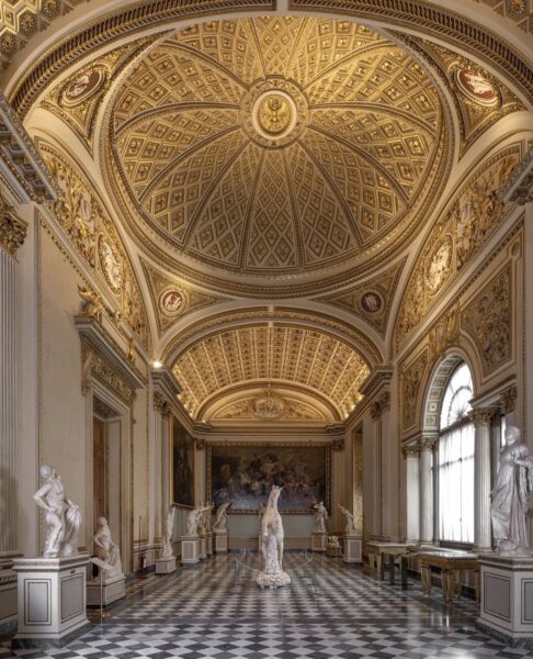 Bogato zdobiona sala w galerii Uffizi we Florencji, z marmurowymi rzeźbami ustawionymi wzdłuż ścian, dużym obrazem na końcu sali oraz imponującym złotym sufitem z geometrycznym i roślinnym ornamentem. Podłoga w biało-czarną szachownicę, wysokie okna po prawej stronie wpuszczają naturalne światło.