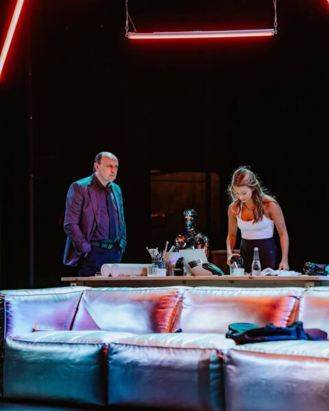 Scena ze spektaklu "Gra" — Julia Wieniawa stoi pochylona nad stołem z butelkami i laptopem, obok niej Adam Woronowicz obserwuje ją uważnie, scenę oświetla neonowe światło, a tło wypełnia nowoczesna scenografia.