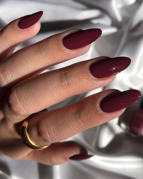 Zdjęcie dłoni ze zbliżeniem na długie, migdałowe paznokcie w kolorze cherry red. Na palcu wskazującym znajduje się masywna, złota obrączka. Dłoń pozuje na tle białego materiału.