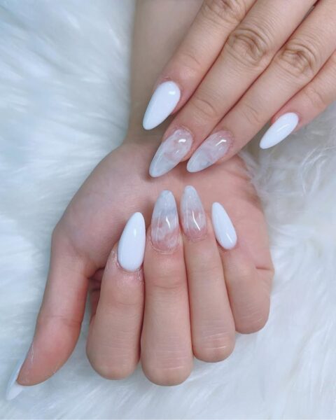 Zdjęcie dłoni z długimi, migdałowymi paznokciami pomalowanymi na biało i ozdobione wzorem marmurkowym. Dłonie układają się tak, by prezentować manicure i pozują na tle białego futerka.
