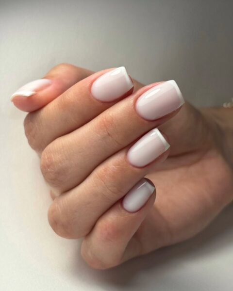 Zdjęcie dłoni ze zbliżeniem na paznokcie, długie i kwadratowe. Są pomalowane w stylu french manicure dwoma odcieniami bieli: kredowej oraz kremowej. Dłoń pozuje na białym blacie.