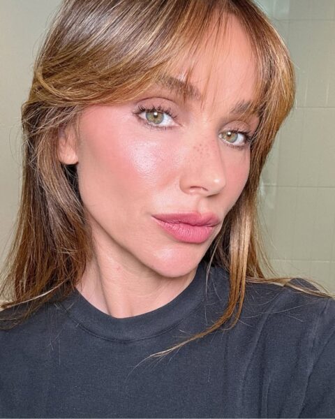 Zdjęcie kobiety, która stoi w łazience i robi sobie selfie. Ma długie, brązowe włosy rozpuszczone na plecy i grzywkę curtain bangs zaczesaną na boki. Ubrana jest w czarną bluzkę.