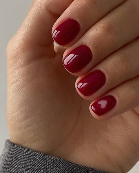 Zdjęcie dłoni z krótkimi, owalnymi, ciemnoczerwonymi paznokciami ze wzorem w kształcie serca w stylu negative space na paznokciu małego palca.