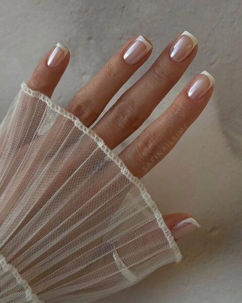 Zdjęcie dłoni ze zbliżeniem na długie, kwadratowe paznokcie w stylu french manicure z perłowym wykończeniem. Widać fragment białego, tiulowego rękawa.