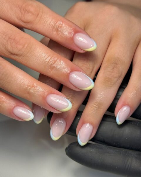 Zdjęcie dłoni z długimi, owalnymi paznokciami z modnym french manicure na koniec czerwca 2025. Są pomalowane na kolor nude z żółto-niebieską końcówką. Dłonie pozują na tle białego blatu.