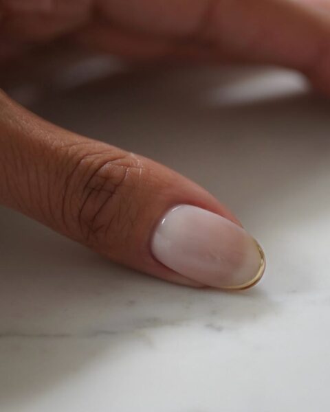 Zdjęcie dłoni ze zbliżeniem na kciuk. Jest pomalowany w stylu french manicure w kolorze mlecznej bieli ze złotą koncówką. Dłoń pozuje na tle marmurowego blatu.