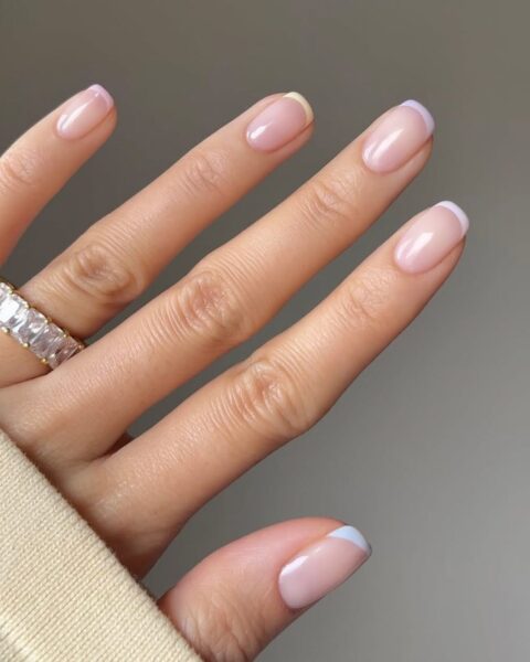 Zdjęcie dłoni z długimi, kwadratowymi paznokciami pomalowanymi w stylu french manicure z pastelowymi końcówkami. Na palcu serdecznym widać masywny pierścień z kryształkami.