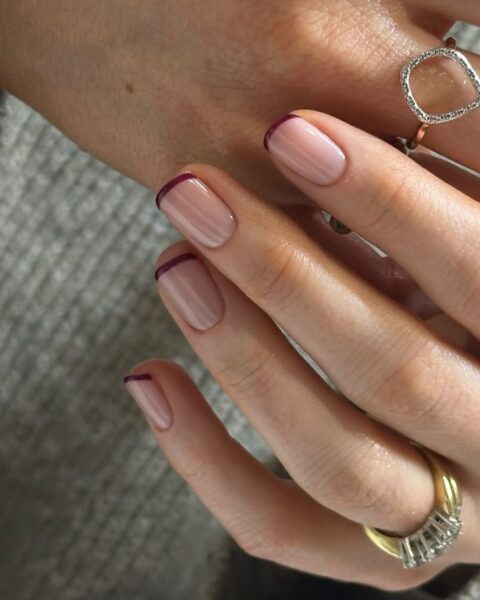 Zdjęcie dłoni z długimi, kwadratowymi paznokciami. Są pomalowane w stylu french manicure z końcówką w kolorze czerwieni cherry red. Na palcach widać pierścionki. Dłonie pozują tak, by prezentować manicure.