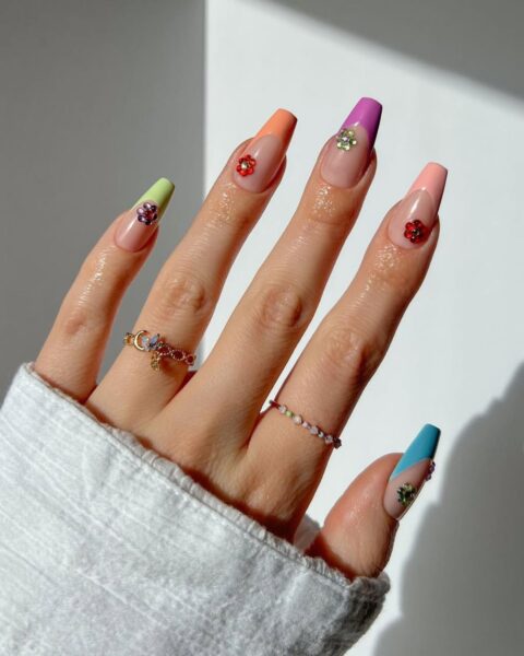 Zdjęcie dłoni z długimi, kwadratowymi paznokciami. Są pomalowane w stylu french manicure, gdzie każdy paznokieć ma inną końcówkę, a do tego paznokcie zdobią kryształki 3D. Na palcach widać kilka pierścionków z kryształkami.