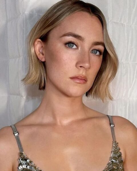 Zdjęcie kobiety w krótkich, blond włosach z fryzurą w stylu jawline bob. To zdjęcie portretowe. Ma odsłonięte ramiona, ale widać ramiączka cekinowego topu. Pozuje na białym tle.