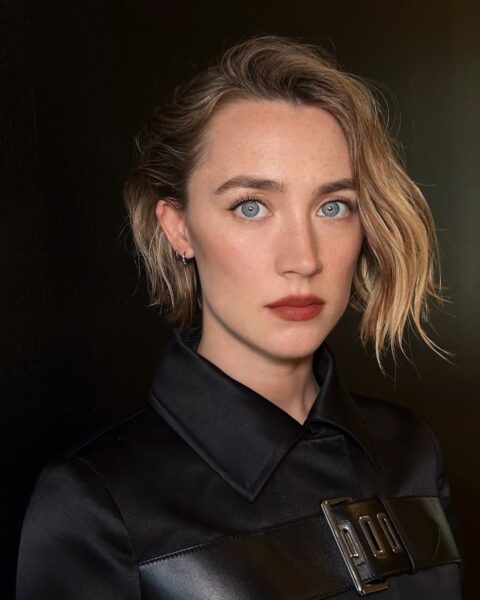 Zdjęcie aktorki Saoirse Ronan w krótkich, blond włosach typu bob. Ubrana jest w czarną koszulę z czarnymi wstawkami. Pozuje na tle czarnej ściany.