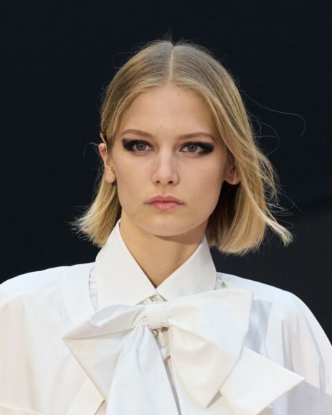 Zdjęcie portretowe modelki, która ma blond włosy ścięte na boba, luźno zaczesane. Ma mocny, czarny makijaż oczu. Ubrana jest w białą koszulę z efektowną kokardą.