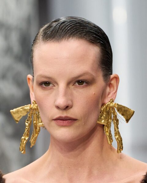 Zdjęcie portretowe modelki. Jej fryzura na wesele na krótkie włosy jest zaczesana na gładko i wykończona w stylu wet look. Ma do tego masywne, złote kolczyki. Ubrana jest w sukienkę z dekoltem karo.