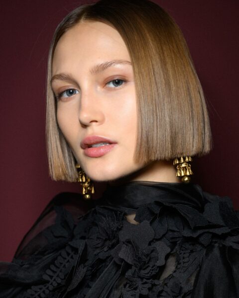 Zdjęcie portretowe modelki, która ma krótkie włosy typu bob w kolorze ciemnego blondu. Są idealnie gładkie i lśniące, a linia cięcia bardzo równa. Kobieta ubrana jest w czarną bluzkę z falbankami i kwiatami. Spod włosów wystają długie, złote kolczyki.