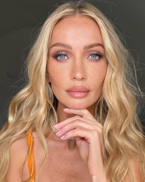 Zdjęcie dojrzałej kobiety w długich, blond włosach ułożonych w stylu beachy waves. Ma delikatny makijaż twarzy. Ubrana jest w pomarańczowy top, a na szyi ma złoty chooker. Podpiera brodę o rękę.