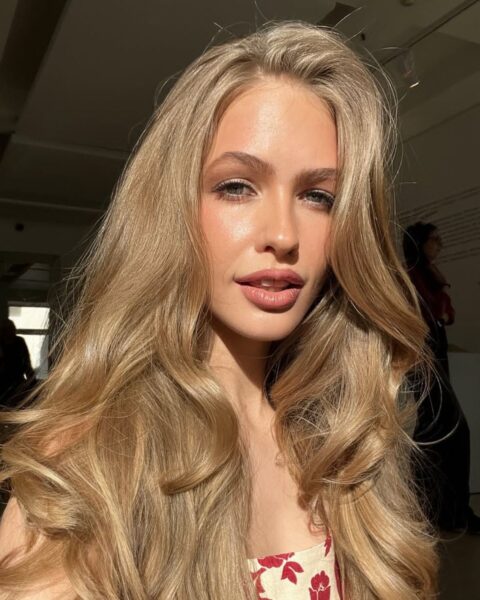 Zdjęcie kobiety w blond włosach, długich, falowanych, odbitych od nasady. Ubrana jest w kremową sukienkę z nadrukiem czerwonych kwiatów. Robi sobie selfie, ma twarz oświetloną słońcem.