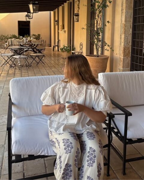 Zdjęcie kobiety siedzącej na patio. Siedzi na fotelu, pije kawę z kubka, obraca się do tyłu. Ubrana jest w bluzkę z rękawem motylkowym, który jest sposobem na to, jak zamaskować grube ramiona. Do tego ma białe spodnie w błękitne kwiaty. Ma długie, ciemnoblond włosy rozpuszczone na ramiona.