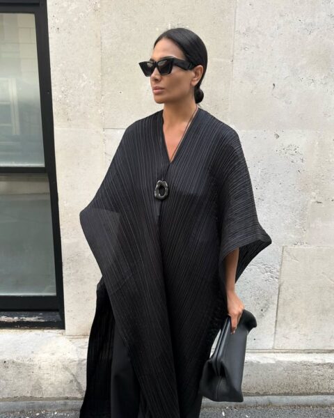 Zdjęcie kobiety ubranej w czarna sukienkę maxi o kroju kimono z długimi rękawami. Ma ciemne włosy związane gładko w kok z tyłu. Na nosie ma okulary przeciwsłoneczne. W ręce niesie czarną torebkę. Stoi na ulicy i patrzy w dal.