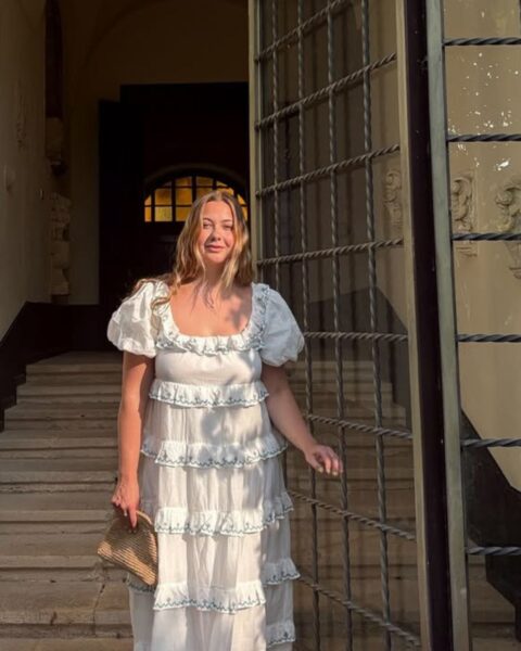 Zdjęcie kobiety z długimi, blond włosami. Jest ubrana w sukienkę maxi o kroju empire z falbankami i rękawami z bufkami, które pomagają zamaskować szerokie ramiona. Stoi w bramie do starej kamienicy.