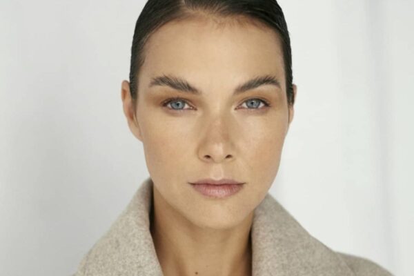 Zdjęcie kobiety, brunetki, która wiedziała, jak używać korektora, aby odmłodzić spojrzenie. Ma gładką cerę i naturalny look. Ma ciemne, gładko związane włosy. Pozuje przodem, ubrana w beżową marynarkę.