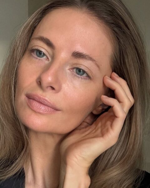 Zdjęcie kobiety, blondynki, w naturalnym, lekkim makijażu, który zasłania jej niedoskonałości, cienie pod oczami i wyrównuje koloryt. Patrzy w bok i przykłada rękę do twarzy.