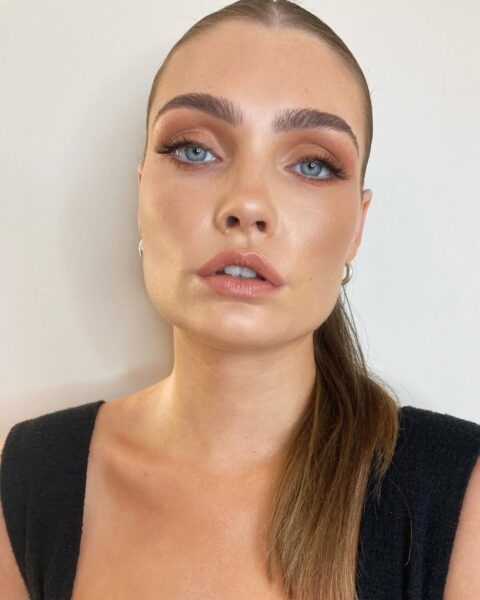 Zdjęcie kobiety w ciemnoblond włosach, związanych gładko z tyłu. Ubrana jest w czarny top bez rękawów. Ma makijaż w stylu glamour i pozuje na wprost obiektywu tak, by prezentować twarz.