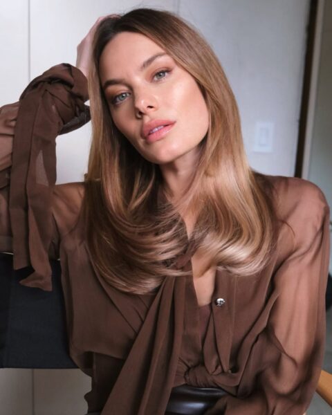 Zdjęcie dojrzałej kobiety, Camille rowe, która ma nowy, modny kolor włosów na lato 2025: light suede blonde z mocnym połyskiem. Włosy są cieniowane, podwinięte na końcach, sięgają poniżej ramion. Kobieta ubrana jest w brązową bluzkę, siedzi na krześle i podpiera głowę o rękę.