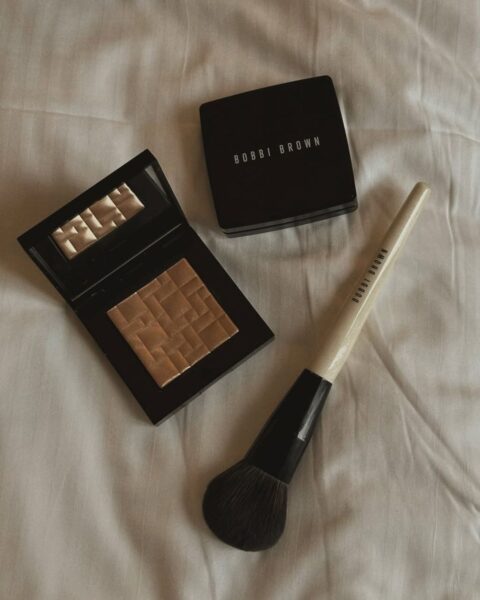 Zdjęcie rozświetlacza w czarnej kasetce marki Bobbi Brown. Obok leży pędzel tej samej marki. Wszystko leży na białej pościeli.