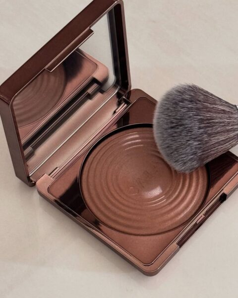 Zdjęcie bronzera do konturowania w formie prasowanej w brązowej kasetce. Produkt ma lusterko i tłoczenie w bronzerze. Leży na nim pędzel do konturowania.