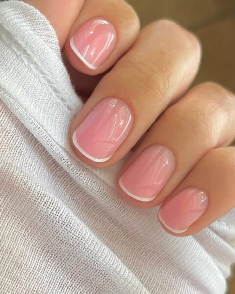 Zdjęcie dłoni z krótkimi, kwadratowymi paznokciami ozdobione stylizacją w stylu micro french manicure z bardzo cienkim, białym paskiem przy brzegach paznokci. Dłoń pozuje tak, by prezentować manicure.