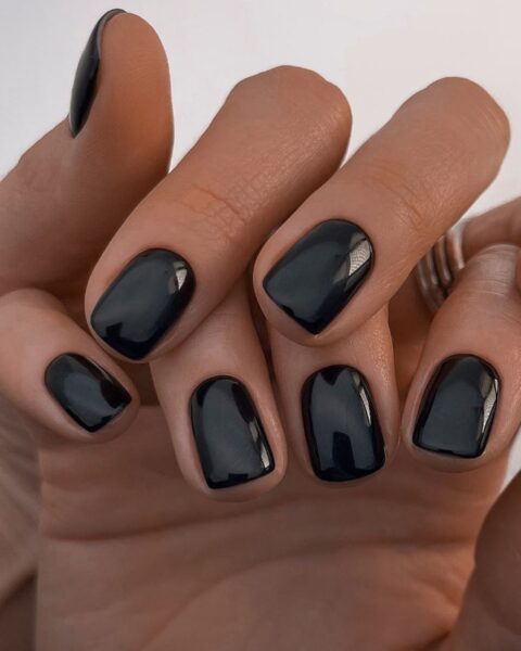 Zdjęcie dłoni ze zbliżeniem na krótkie, kwadratowe paznokcie. Są pomalowane na czarno. Dłonie są splecione, w taki sposób pozują, by prezentować manicure.