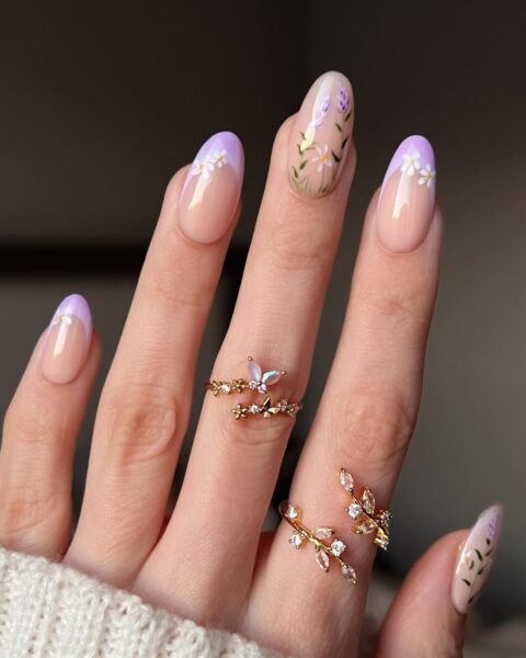 Zdjęcie dłoni z długimi, migdałowymi paznokciami z french manicure w kolorze lawendy, ozdobione wzorem kwiatów. Na palcach znajdują się złote pierścionki z kryształkami i motywami liści.