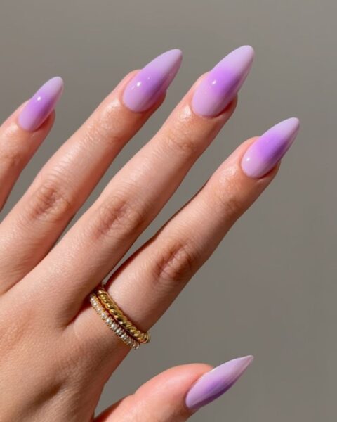 Zdjęcie dłoni z długimi, migdałowymi paznokciami w kolorze lawendy i ozdobione wzorem w stylu aura nails. Na palcu wskazującym znajduje się podwójny pierścionek w kolorze złoto-srebrnym.