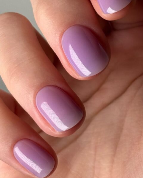 Zdjęcie dłoni z mocnym zbliżeniem na krótkie, owalne paznokcie. Są pomalowane na lawendowo i tworzą stylizację lavender donut glaze z mocnym połyskiem.
