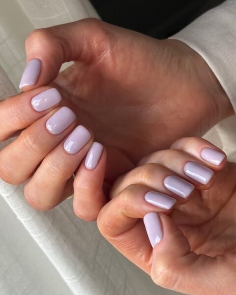 Zdjęcie dłoni z krótkimi, kwadratowymi paznokciami pomalowanymi na kolor lawendy z mlecznym wykończeniem, czyli milk lavender manicure. Dłonie pozują tak, by prezentować manicure przy zasłonie okiennej.