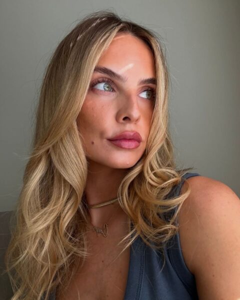 Zdjęcie kobiety, która zrobiła sobie selfie od dołu. Ma długie, blond włosy, patrzy w bok i na jej twarz padają promienie słońca. Ma makijaż toasty glam, czyli ocieplający i rozświetlający. Ubrana jest w ciemnoszary top bez rękawów.