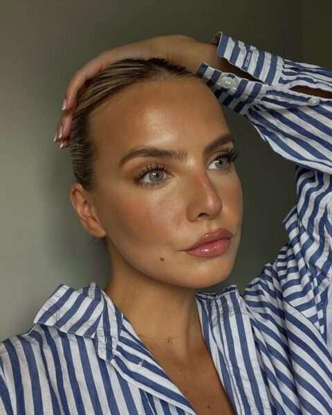 Zdjęcie kobiety w blond włosach zaczesanych do tyłu. Ma ciepły, rozświetlający makijaż w kolorach brązu i koralu. Ubrana jest w białą koszulę w niebieskie paski. Opiera rękę o głowę. Patrzy w bok, gdzieś w dal.