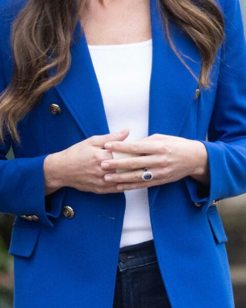 Zdjęcie ze zbliżeniem na dłonie i paznokcie Kate Middleton. Księżna jest ubrana w białą bluzkę, czarne spodnie i kobaltową marynarkę.