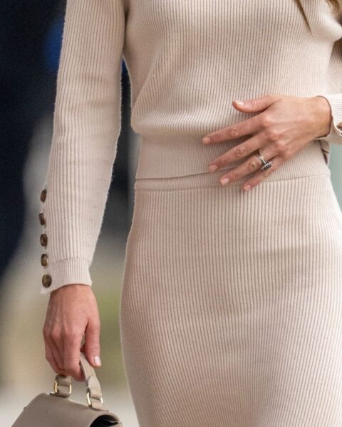 Zdjęcie ze zbliżeniem na manicure Kate Middleton. Księżna jest ubrana w dzianinowy, beżowy sweter z guzikami przy rękawach oraz ołówkową spódnicę beżową z dzianiny. W ręce trzyma torebkę.
