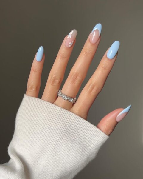 Zdjęcie dłoni ze zbliżeniem na długie, owalne paznokcie z niebieskim manicure. Jeden paznokieć jest ozdobiony wzorem kwiatów. Na środkowym palcu znajduje się pierścionek z kryształami.