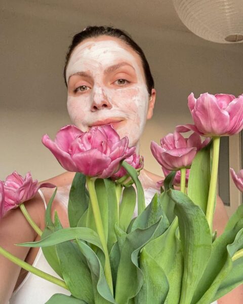 Zdjęcie kobiety w maseczce na twarzy. Jest w trakcie pielęgnacji marry me skin przygotowującej jej cerę do ślubu. Ma ciemne włosy związane z tyłu. Ubrana jest w biały top na ramiączkach. Przed sobą trzyma duży bukiet różowych tulipanów.