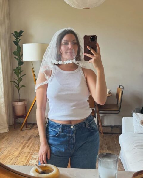 Zdjęcie kobiety, która robi sobie selfie w lustrze. Jest ubrana w biały top, niebieskie jeansy, a na głowie ma wianek ślubny.