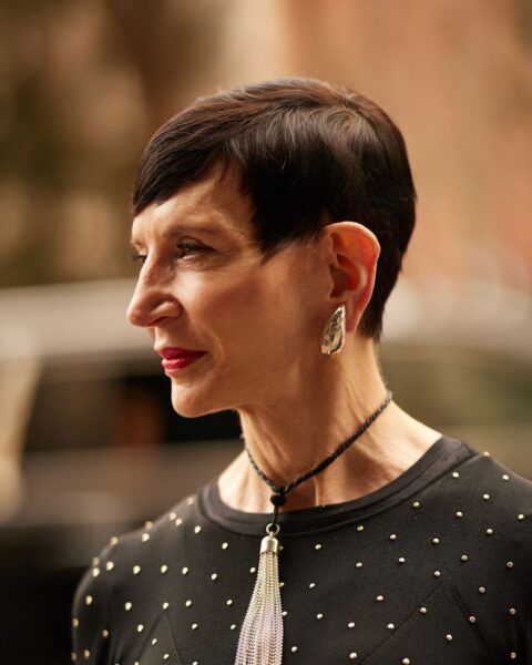 Zdjęcie dojrzałej kobiety w krótkich, ciemnych włosach ściętych w stylu pixie cut. Ma na twarzy najlepszy podkład na lato dla cery dojrzałej. Ubrana jest w czarną bluzkę w groszki, na szyi ma naszyjnik z chwostem oraz masywne kolczyki w uszach.