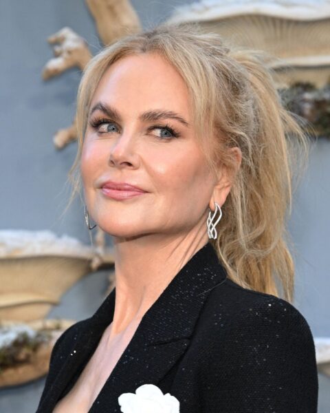 Zdjęcie aktorki Nicole Kidman. Ma blond włosy związane w koński ogon, z kosmykami przy twarzy. Ubrana jest w czarną marynarkę. W uszach ma srebrne kolczyki. Pozuje na tle niebieskiej, zdobionej ściany.