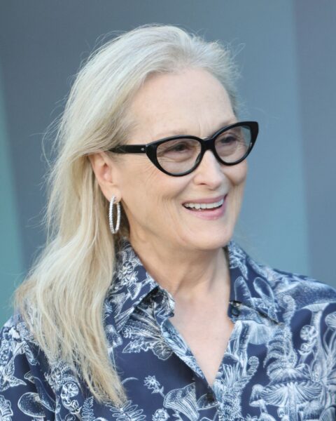 Zdjęcie dojrzałej kobiety, aktorki Meryl Streep. Ubrana jest w granatową koszulę z białym nadrukiem. Ma rozpuszczone, popielate włosy. W uszach wysadzane cyrkoniami kolczyki kółka. Na nosie ma okulary przeciwsłoneczne w kolorze czarnym.